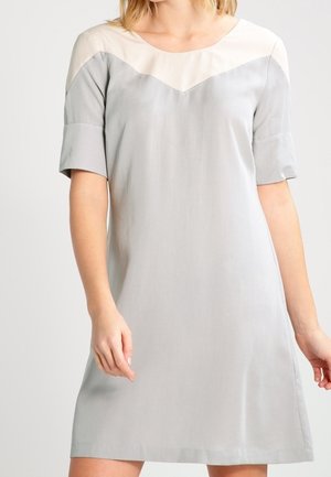 Vrouw die een lichtgrijze jurk met korte mouwen, een beige juk-detail en een ronde halslijn draagt, staand tegen een effen achtergrond.
