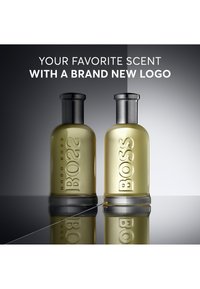 Twee Boss-geurflesjes naast elkaar op een reflecterend oppervlak; links met oud logo, rechts met nieuw logo; tekst ter promotie van het nieuwe logo erboven.