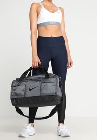 Szara torba sportowa z czarnymi akcentami, z logo Nike, wieloma zewnętrznymi kieszeniami i mocnym paskiem na ramię. Idealna do użytku sportowego.
