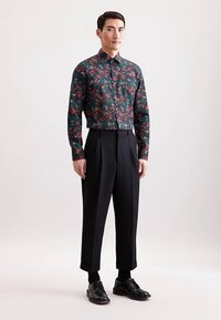 Bloemenoverhemd met knopen in donkergroen, met rode en gele patronen. Gecombineerd met zwarte geplooide broek en zwarte nette schoenen.