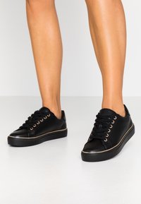 Black leather sneakers featuring a round toe, low top design, eight lace eyelets, and a contrasting gold trim along the sole.