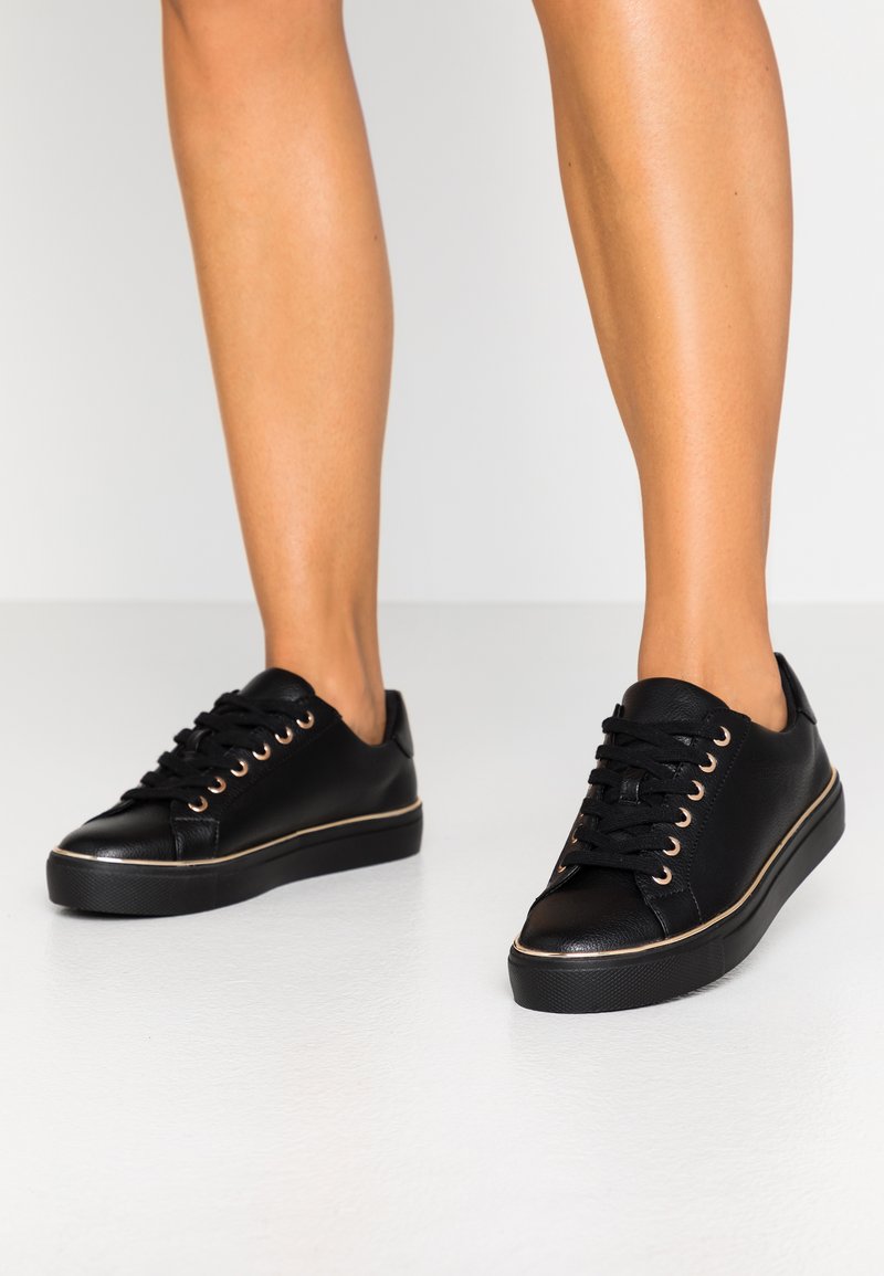 Black leather sneakers featuring a round toe, low top design, eight lace eyelets, and a contrasting gold trim along the sole.