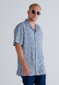 Chemise à manches courtes à boutons en bleu clair avec un motif géométrique. Fabriquée dans un tissu à la texture douce, comportant des boutons blancs et une coupe décontractée.