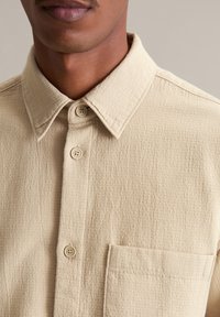 Beige overhemd met knopen, gemaakt van textuurstof, met een puntige kraag, een borstzak en vier bijpassende knopen op de sluiting.