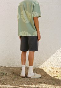 Grünes Kurzarmhemd mit großem hellgrünem Blattmuster, kombiniert mit dunklen Shorts. Cremefarbene Sandalen, weiße Socken, stehend auf Erdoberfläche.