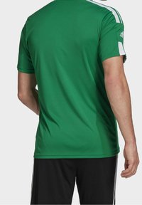 Man som bär en grön Adidas sporttröja med vita ränder på ärmarna och svarta shorts med vita sidränder, sedd bakifrån.