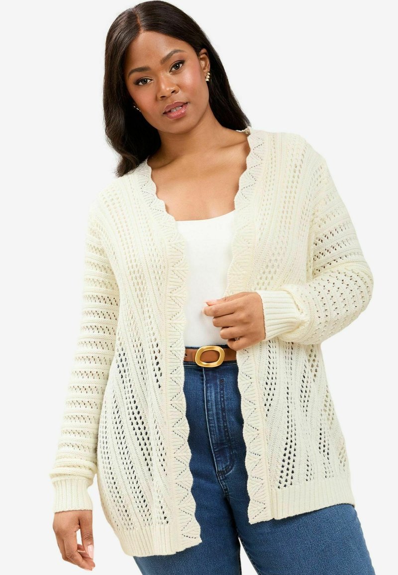 Cardigan crema de punto abierto con bordes festoneados y puños acanalados, superpuesto a una blusa blanca, combinado con jeans de tiro alto azules y un cinturón marrón.