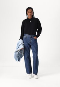 Sudadera negra con logo blanco, jeans de mezclilla azul de cintura alta y zapatillas blancas. Sosteniendo una chaqueta azul clara con acentos coloridos.