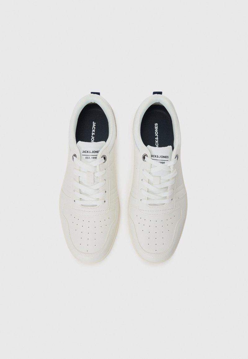 Des baskets blanches avec des détails perforés et une doublure intérieure noire, dotées de lacets et d'un logo sur la languette, sur un fond uni.