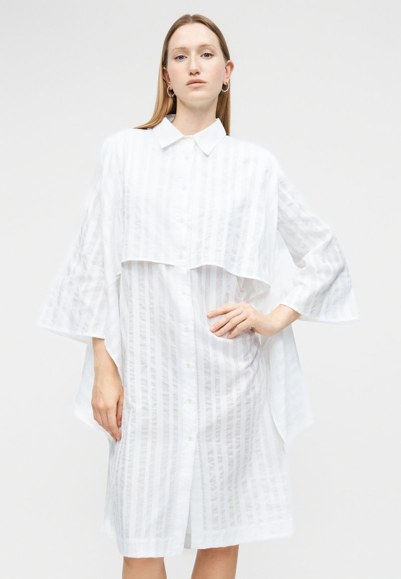 Femme aux cheveux blonds portant une robe chemise blanche transparente à rayures avec des manches larges, debout avec une main sur la hanche devant un fond uni.