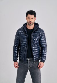 Marineblauwe pufferjas met glanzende afwerking, ritssluiting aan de voorkant en capuchon. Voorzien van geribbelde manchetten en een borstzak. Draagt over een zwart shirt en een jeans.