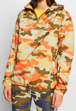 Osoba ubrana w wielokolorową kurtkę przeciwwiatrową w kamuflażu o odcieniach pomarańczowym, beżowym i brązowym oraz dopasowane legginsy w kamuflażu.