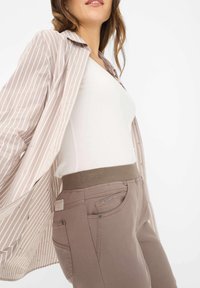 Camicia a righe beige chiaro con vestibilità ampia, indossata sopra un top bianco e pantaloni elasticizzati marroni con vita a costine e tasca con zip.