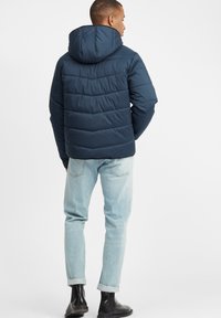 Navy puffer jacket with a hood, featuring horizontal stitching, smooth texture, and cuffed sleeves; paired with light denim jeans and black boots.