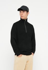 Pull à col roulé noir côtelé avec un col zippé mi-hauteur, associé à un pantalon beige clair. Le tissu semble doux et texturé.