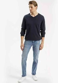 Granatowy sweter w serek, lekki materiał, zestawiony z jasnoniebieskimi dżinsami. Model ma na sobie białe trampki, prezentując swobodny, dopasowany styl.