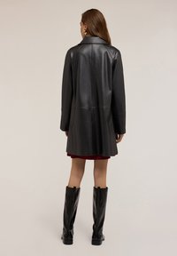 Kobieta w czarnym skórzanym płaszczu, czarnych wysokich butach z ćwiekami i czerwonej spódnicy, stojąca tyłem na jednolitym tle.