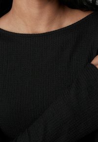 Schwarzes geripptes Strickoberteil mit weitem Ausschnitt und langen Ärmeln. Die Textur ist weich und leicht erhaben, was die Dicke des Stoffes betont.