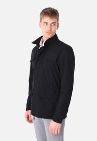 Giacca nera con colletto alto, molteplici tasche frontali e chiusura con zip. Il tessuto appare testurizzato e resistente, adatto per un uso all'aperto.