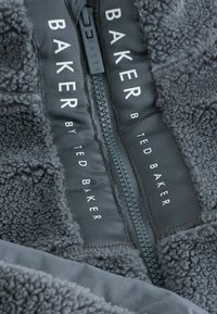 Szara kurtka polarowa z widocznym zamkiem, teksturowanym materiałem oraz białymi metkami "BAKER by TED BAKER" na kontrastowym czarnym tle.