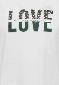 T-shirt blanc avec le mot "LOVE" en vert et un motif léopard, avec des détails argentés et le texte "All you need is" en dessous.