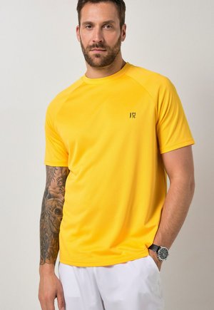 Mann mit tätowiertem linkem Arm, der ein gelbes Kurzarmhemd mit kleinem Logo und weiße Hose trägt, steht vor einem einfarbigen Hintergrund.