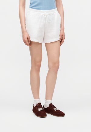 Femeie purtând pantaloni scurți albi cu șnur, șosete albe și adidași bordo închis cu șireturi, stând în fața unui fundal simplu, deschis.