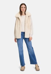 Cremefarbene Kunstpelzweste mit strukturiertem Kragen, über einem hellen Oberteil und blauen weiten Jeans getragen. Beigefarbene Slipper mit dicker Sohle.