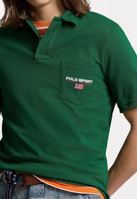 Personne portant un polo vert Polo Sport Ralph Lauren avec un logo sur la poche et une chemise rayée orange en dessous.
