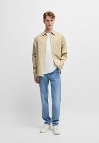 Camicia beige con bottoni, due tasche sul petto, indossata sopra una maglietta bianca, abbinata a jeans azzurri chiari e sneakers bianche. Design semplice, vestibilità casual.