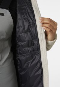 Een geborduurde beige jas staat open en onthult een gladde, donker navy binnenvoering met meerdere kleine zakken; een grijze textuur kledingstuk is gedeeltelijk zichtbaar daaronder.