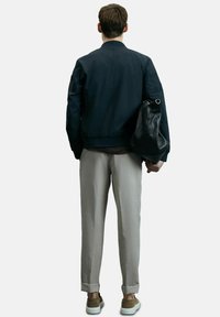 Mann, der von hinten steht, trägt eine schwarze Jacke, hellgraue hochgekrempelte Hosen, braune Sneaker und hält eine schwarze Lederhandtasche in seiner rechten Hand.