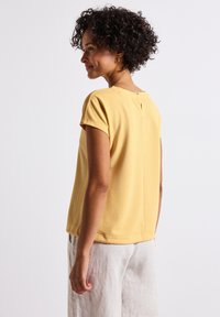 Žlutý texturovaný top s krátkým rukávem a kulatým výstřihem, se zadním detailem ve tvaru klíčové dírky, doplněný volnými kalhotami světelné barvy.