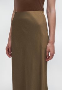 Gros plan d’une personne portant un haut marron ajusté et une jupe taille haute vert olive, les mains détendues le long du corps.