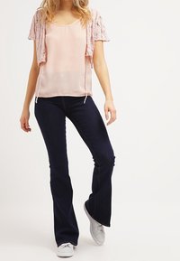 Vaaleanpunainen lyhythihainen neuletakki vaaleanpunaisen paidan päällä, yhdistetty tummansinsisiin leveisiin farkkuihin ja valkoisiin lenkkareihin. Sujuvat kankaat, rento tyyli.