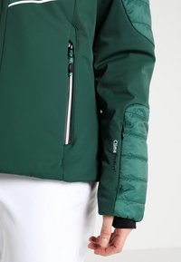 Veste isolante verte avec une surface extérieure lisse, détail matelassé sur les manches, poche zippée et marquage "Clima Protect" sur le poignet.
