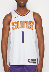 Hombre vistiendo una camiseta de baloncesto blanca de los Phoenix Suns con el número 1 y ribetes en púrpura y naranja, combinada con pantalones cortos morados.