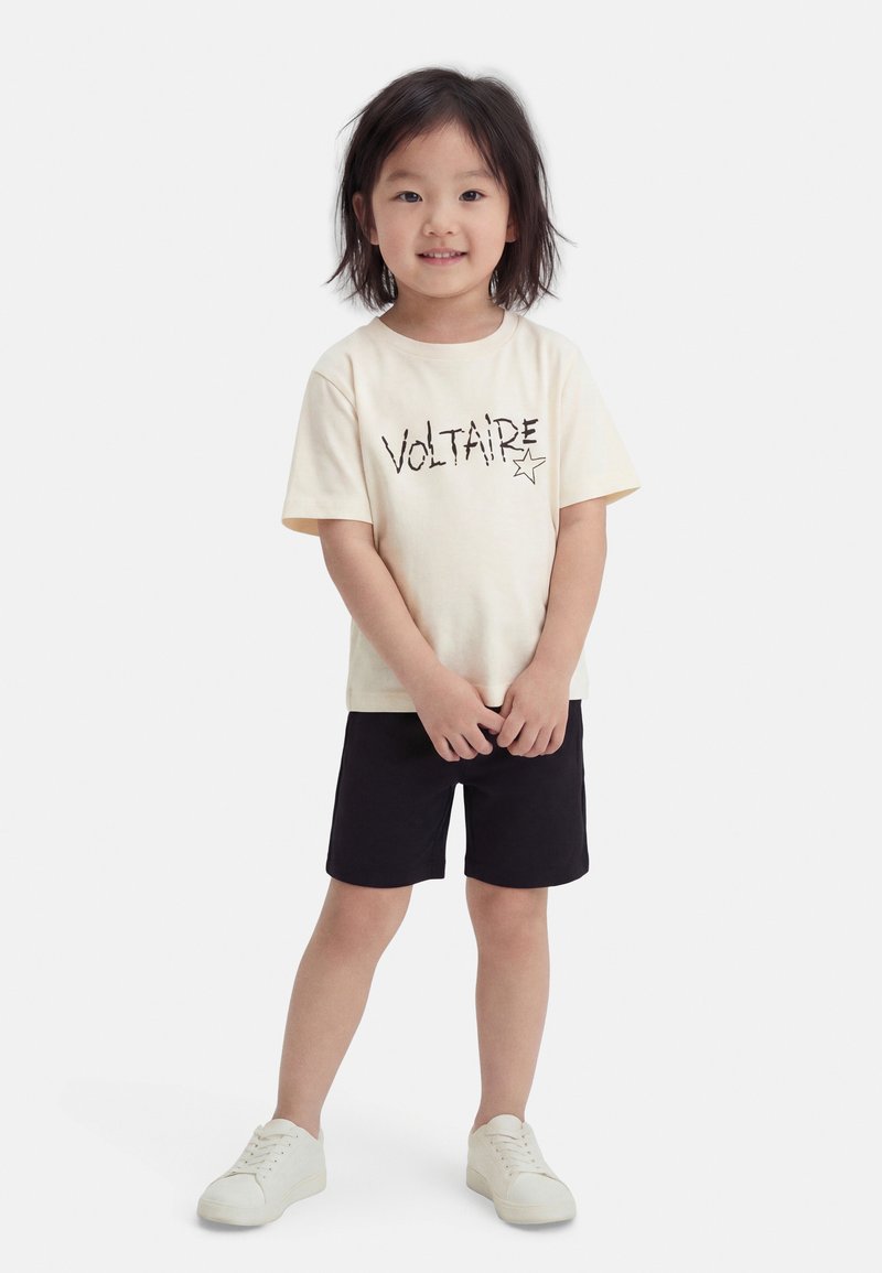 Jeune enfant debout, portant un t-shirt crème "Voltaire", un short noir et des baskets blanches sur un fond blanc uni.