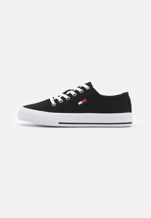 Zapatilla negra de lona de corte bajo con cordones blancos, suela de goma blanca y pequeño logo rojo, blanco y azul en el lateral.