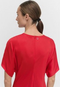 Kobieta z brązowymi włosami spiętymi w kucyk, ubrana w luźną czerwoną bluzkę, ukazana z tyłu na jednolitym jasnym tle.