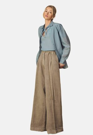 Kobieta ubrana w jasnoniebieską koszulę zapinaną na guziki, na niebieskim dzianinowym topie oraz jasnobeżowe, szerokie spodnie z wysokim stanem i paskiem, stojąca na białym tle.