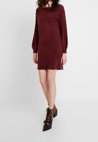 Abito lungo bordeaux a maniche lunghe con scollo rotondo, spacchi laterali e silhouette dritta. Modello che indossa stivaletti alla caviglia in stampa serpente texture.