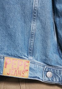 Ljusblå denimjacka med ett märkesläderetikett med rosa text. Den har silverfärgade detaljer och synliga sömmar.