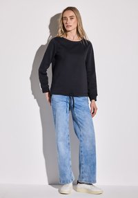 Sudadera negra de cuello redondo con mangas largas, combinada con jeans de pierna ancha de color azul claro y zapatillas blancas. Ajuste casual. Detalle de cordón en la cintura.