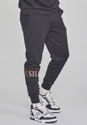 Persona con pantalones jogger negros con texto dorado en una pierna y zapatillas blancas de caña alta con patrones negros y calcetines blancos gruesos.