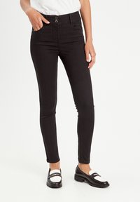Jean skinny noir taille haute, avec deux boutons à l'avant, quatre poches et un tissu en denim lisse et extensible. Associé à des mocassins noir et blanc.
