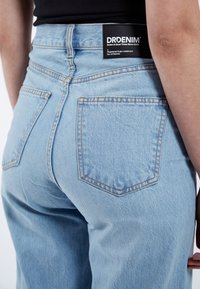 Jeans de tiro alto azul claro con textura suave, con bolsillos traseros y costuras en contraste. Etiqueta negra en la cintura.
