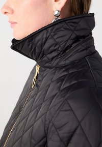 Zwarte gestikte jas met een hoge kraag en een gouden rits. Glad, glanzende textuur met geometrische stikpatronen. Metaalachtige studs als oorbellen.