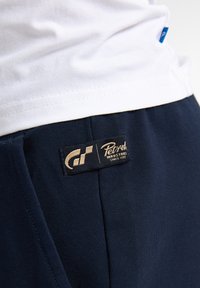 Námořnické modré bavlněné kraťasy s viditelnou černou etiketou s nápisy "Petroci" a "Industries" ve zlatém písmu, doplněné kostkovaným vzorem.