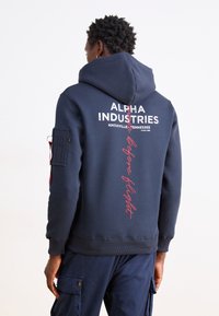 Námornícka modrá mikina s kapucňou s textom "Alpha Industries" v bielej a červenej farbe. Obsahuje bočnú kapsu a elastické manžety a lem. Text na chrbte znie "Pred letom."
